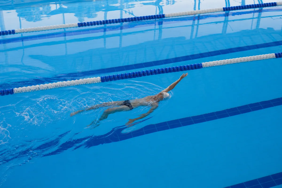 Nageur sur le dos en dos crawlé, visage à l&rsquo;air libre, sous les lignes de plafond d&rsquo;une piscine couverte